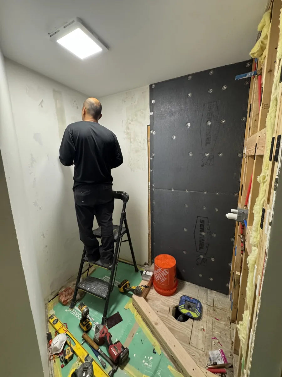 Columbus bathroom remodel in progress: worker on ladder, waterproofing, exposed plumbing, and new LED light fixture.