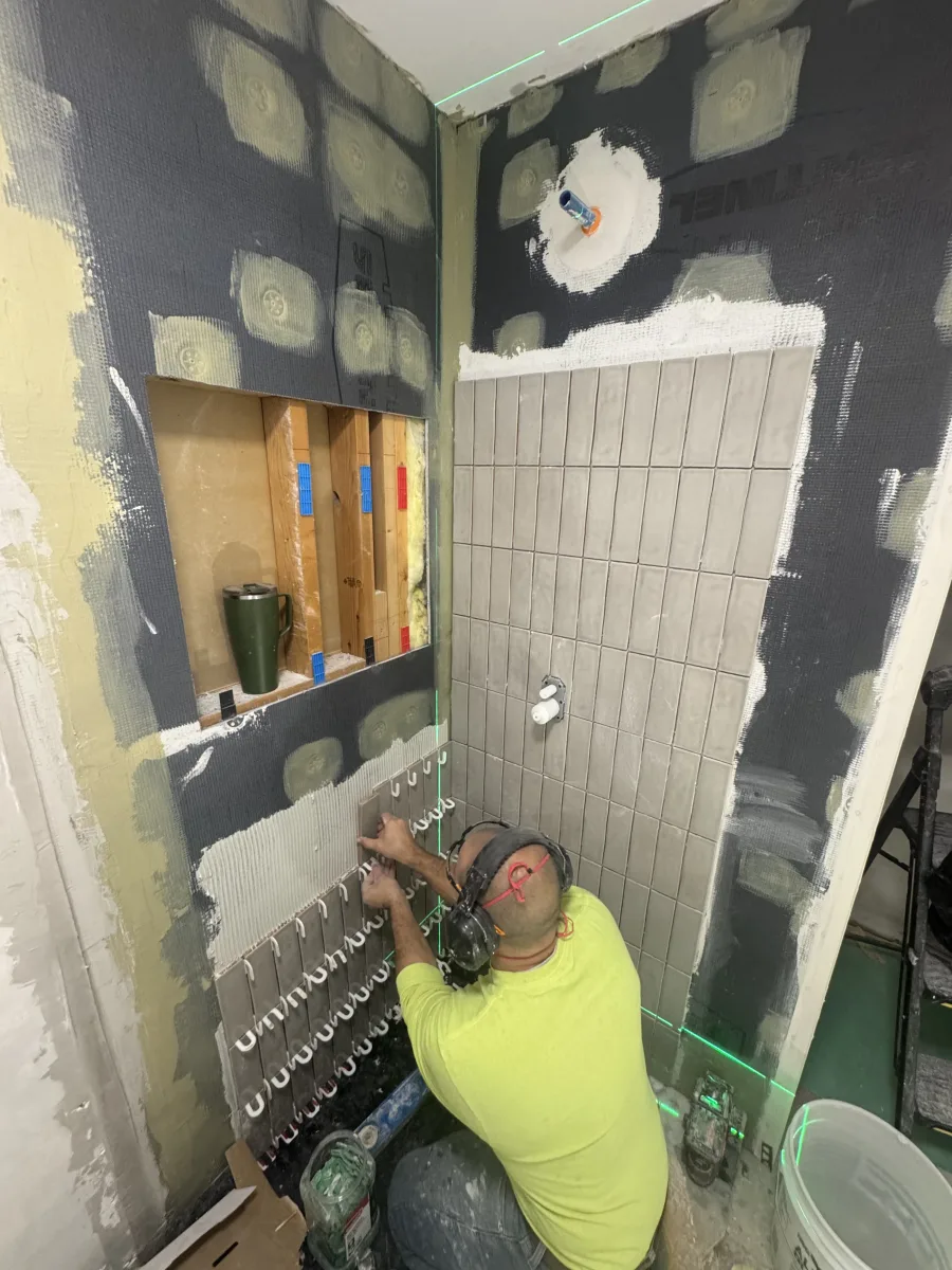 Columbus bathroom remodel: worker installing sleek gray subway tiles in shower with laser level, waterproofing membrane, and wall niche.